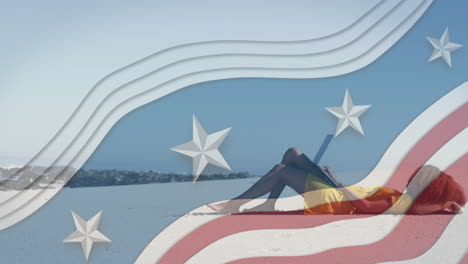 varias estrellas en rayas azules contra una mujer afroamericana usando una computadora portátil acostada en la playa