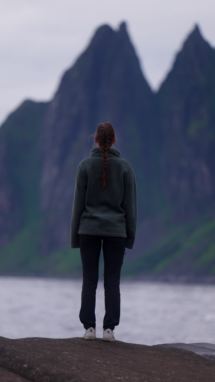 Solo woman stands at Djevelens Tanngard cliffs in Senja, Norway under looming rock formations, vertical telephoto compression