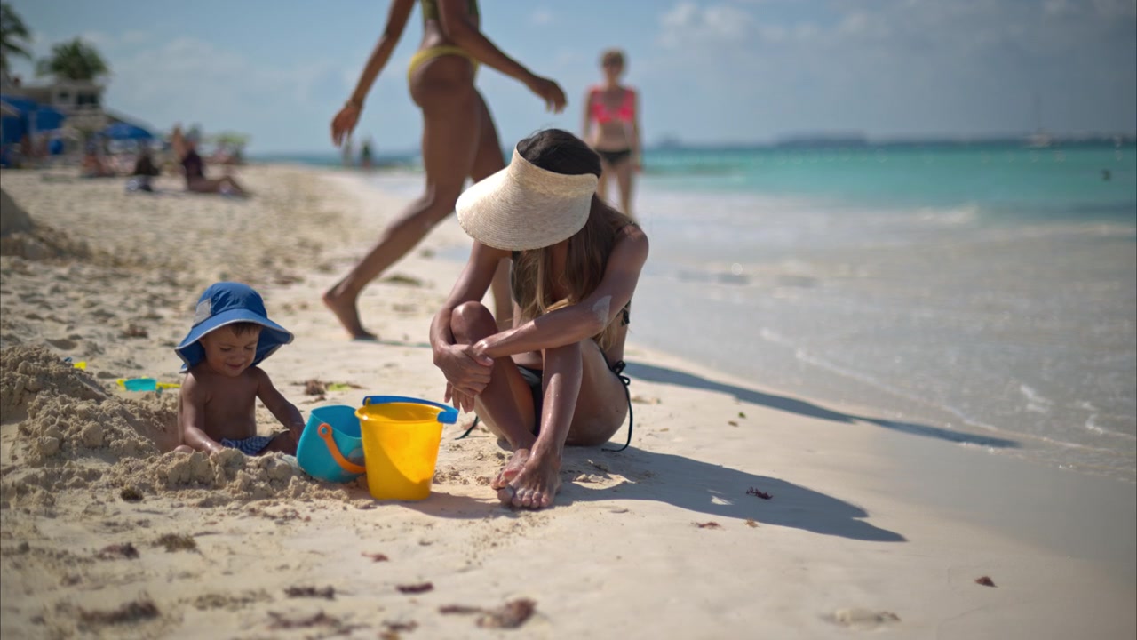 Schattige Spaanstalige peuter speelt op het strand met zand onder toezicht van zijn moeder, terwijl mensen voorbijlopen