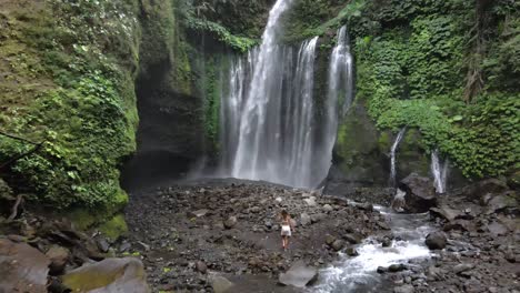 woman walking over rocks towards giant waterfall in rainforest