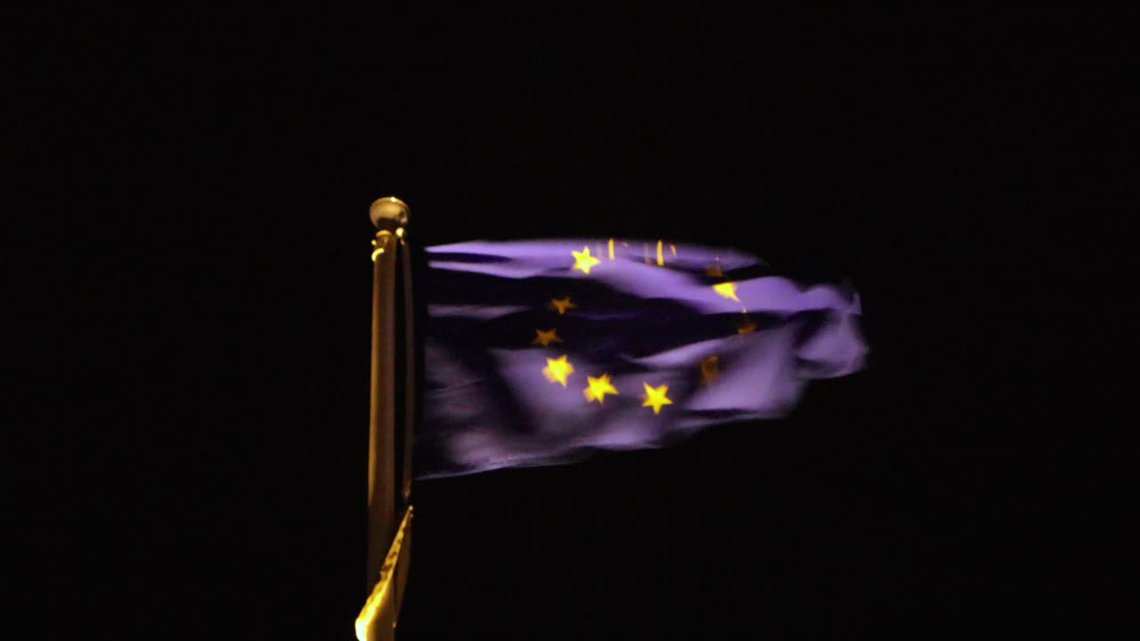Bandiera dell'Unione Europea ben illuminata con stelle dorate che sventolano al vento forte contro un cielo notturno nero profondo.
