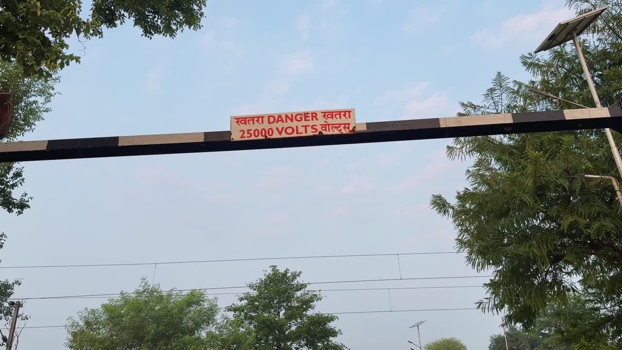 Overhead railway bar with red 25000 volts danger sign against soft morning sky surrounded by green trees as the camera circles around the structure up