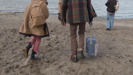 grupo de meninos e meninas adolescentes vestindo roupas de inverno caminhando em direção à praia com mochilas enquanto tiram fotos de seus amigos em um dia nublado