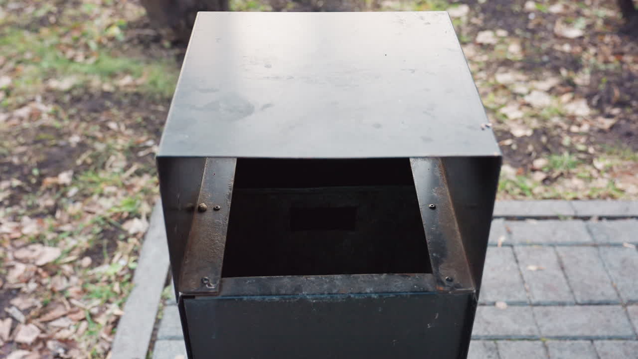 Close up view of public metal trash can placed next to wooden bench in quiet city park, surrounded by leaf covered ground and paved path, highlighting sanitation and urban outdoor cleanliness