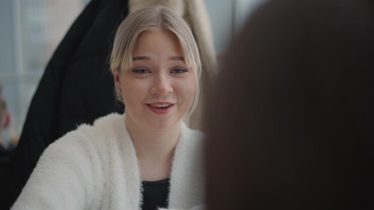 Medium close-up of young woman seated indoors speaking to another person off-camera with slightly open mouth, forward lean, and visible hand gesture that indicate engaged response during conversation