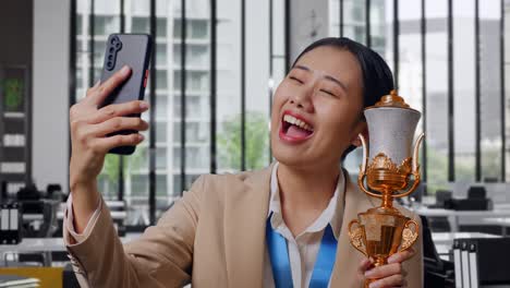 close up of asian business woman in a suit with a gold medal showing a gold trophy to the video call on smartphone in the office