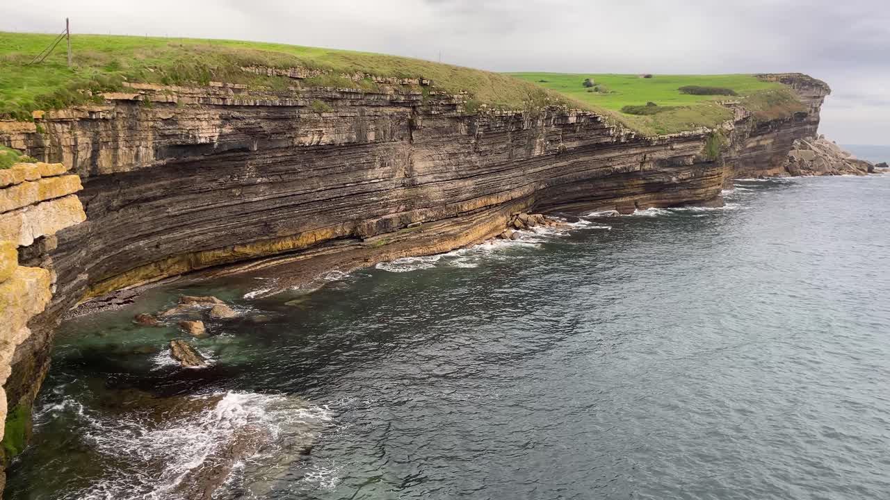 Beautiful cliff of the Cantabrian Sea where we see the sea water crashing against the wall and we appreciate the horizontal strata on the cliff wall, there is a blanket of grass on top.
