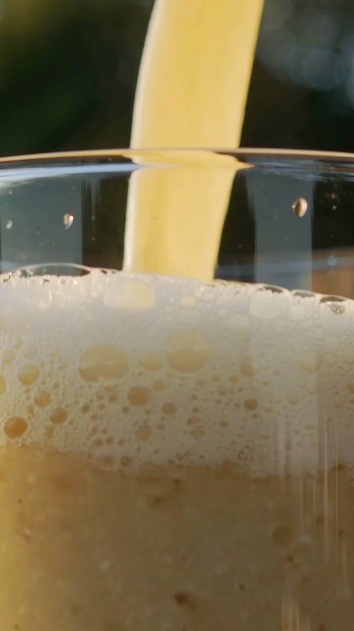 Vertical video: Off-frame pouring starting pale drink filling clear glass, forming foam head for ad