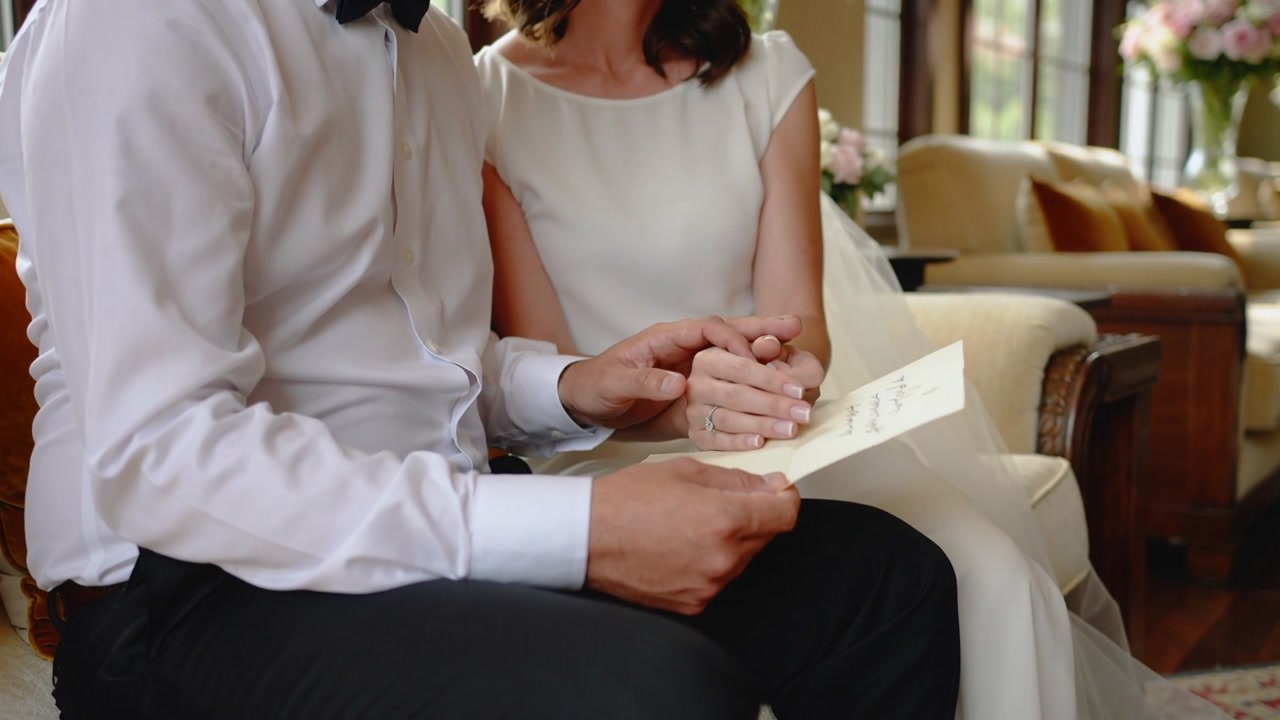 Man opening card, couple in formal attire reading, holding hands on living room sofa, ring visible