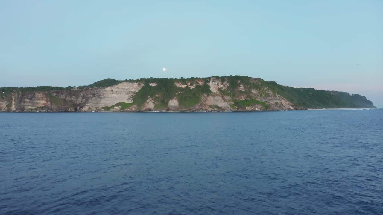 녹색의 울창한 숲으로 뒤덮인 바다 쪽 절벽을 향해 물 위를 날아가는 공중 드론 보기