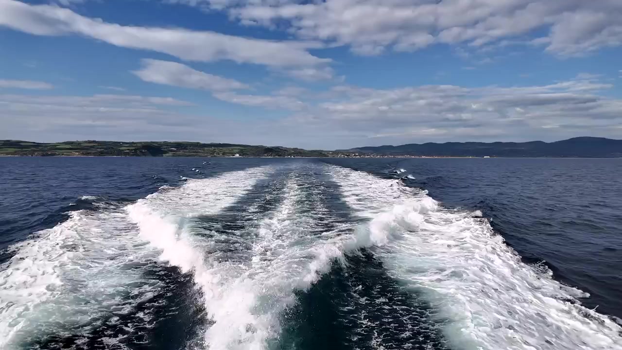 Una barca fende l'acqua blu, lasciando scie bianche nella sua scia. Il sole splende in alto e le nuvole si muovono nel cielo mentre la terra appare lontana. La giornata è limpida e luminosa