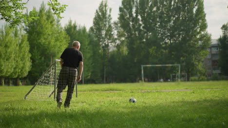 an elderly man kicks a soccer ball near a small goalpost and chases after it as it bounce on the grassy field, a view of a big goal post in the distance