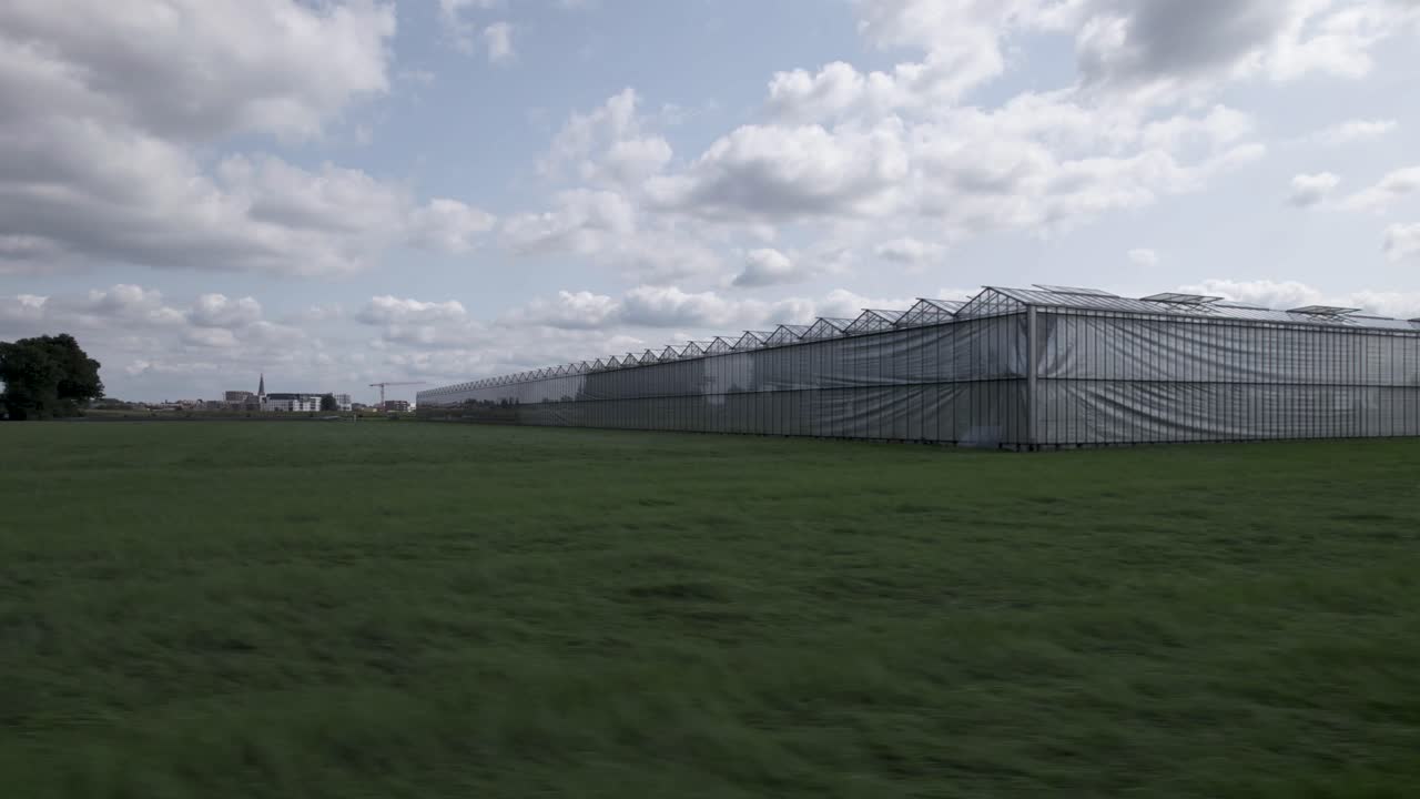 invernadero en el campo de pradera visto desde un lado con reflejo de árboles visibles en el vidrio mientras se acerca a los lados y nubes cumulus en el cielo azul arriba