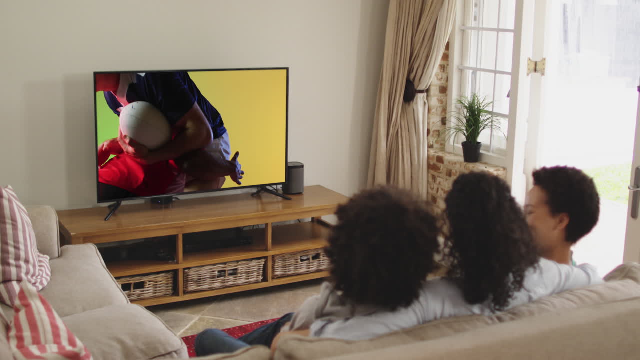 Famiglia birazziale che guarda la TV con diversi giocatori di rugby maschi che giocano sullo schermo.