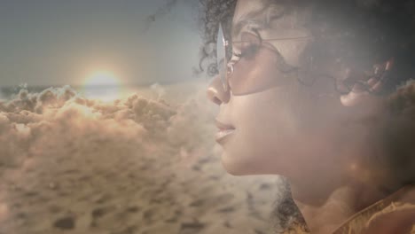 animation of african american woman wearing sunglasses at beach, over clouds