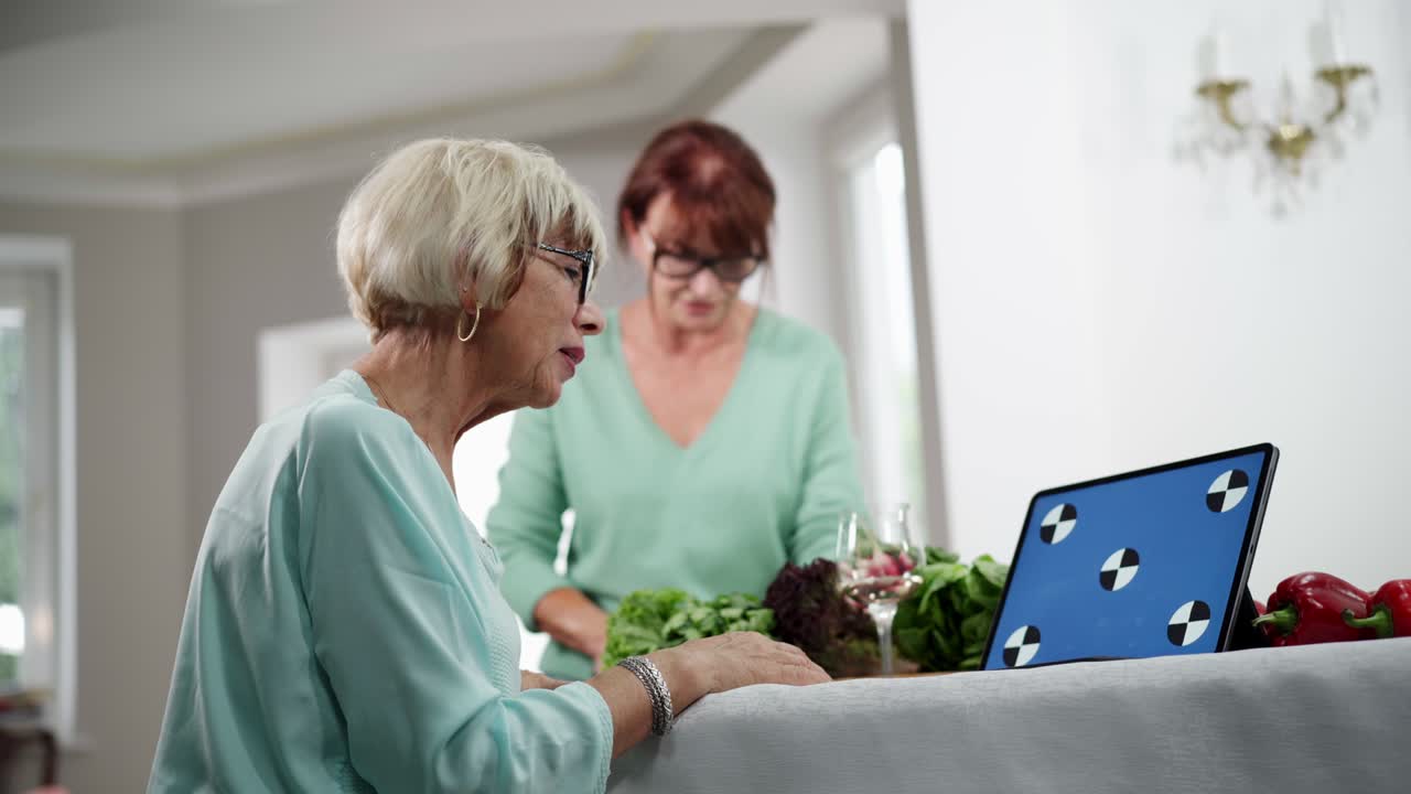 Side view portrait of slim beautiful senior woman reading online recipe on laptop with blue screen as blurred confident friend cutting vegetables at background. Retirees cooking indoors.