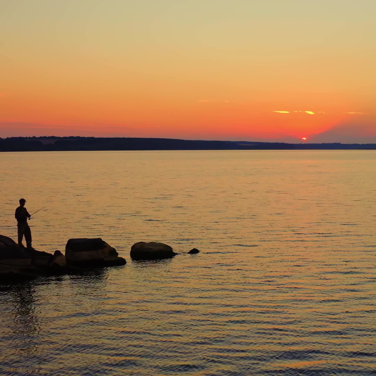 Fisherman at the evening river. Silhouette of a man fishing on the stone in water on the beautiful nature background at sunset.
