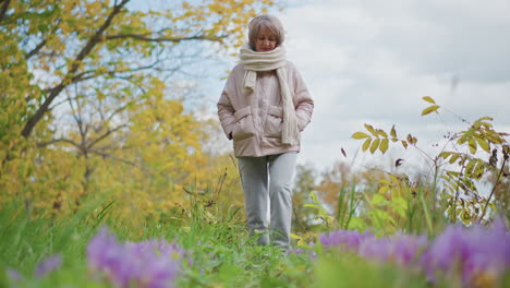 young girl walking thoughtfully through lush autumn garden path holding knitted muffler while dressed in pink coat and grey joggers, surrounded by tall grass, purple blooms and yellowing leaves