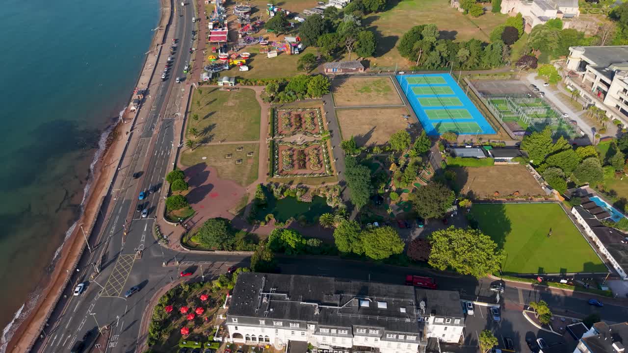 Morning drone flight west over Torquay, UK. Blue sea lies to the left as the camera tilts down, ending above the neat rectangular gardens. Hotels, houses, and greenery frame this seaside scene