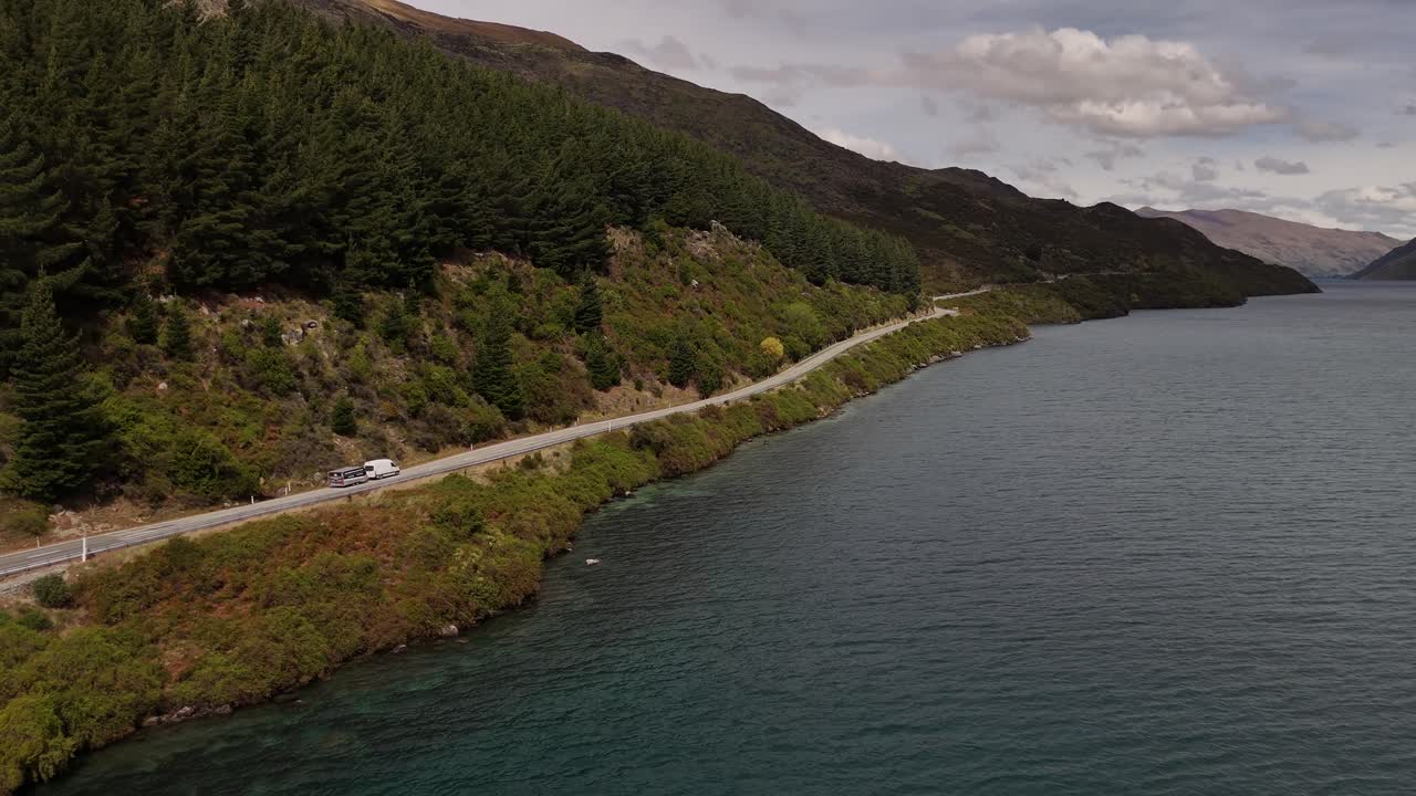 Ripresa aerea in movimento di un furgone che traina un rimorchio lungo una strada panoramica in riva al Lago Wakatipu, nella regione di Otago, Isola del Sud della Nuova Zelanda, circondato da colline boscose sempreverdi.