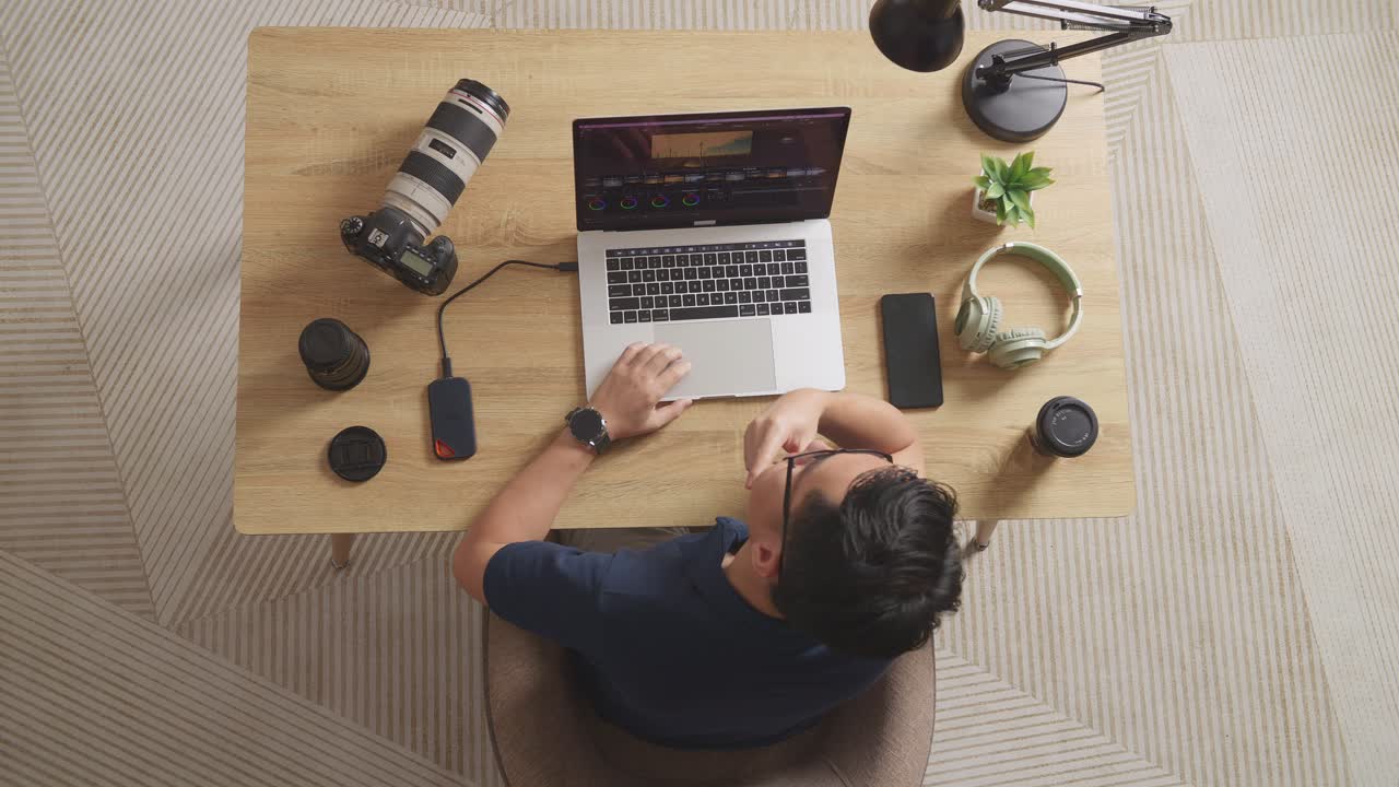 Top View Of Asian Male Color Grading Thinking About The Footage While Sitting In The Workspace Using A Laptop Next To The Camera Editing The Video At Home