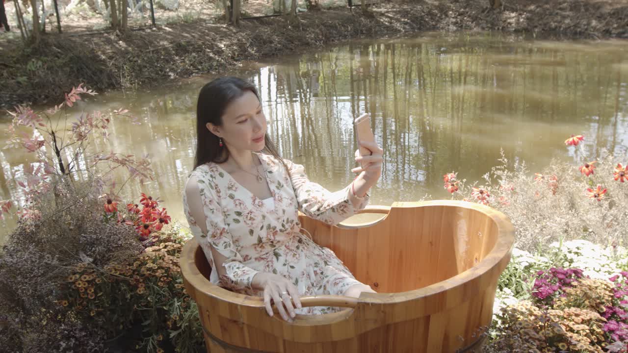 멋진 아시아 여성이 정원 연못 옆의 나무 욕조에 앉아 스마트폰으로 셀카를 찍습니다.