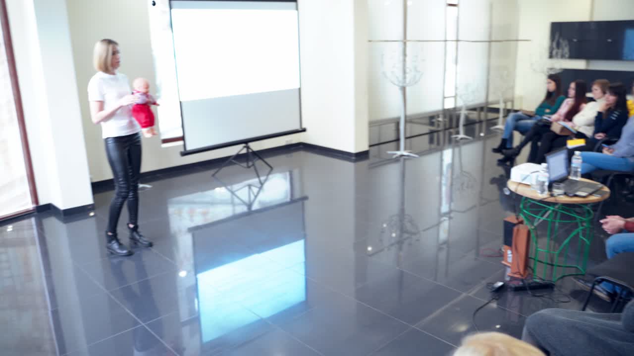Young auditory listening to educator. Woman speaker holding a doll and telling the lecture to listeners in the modern hall.