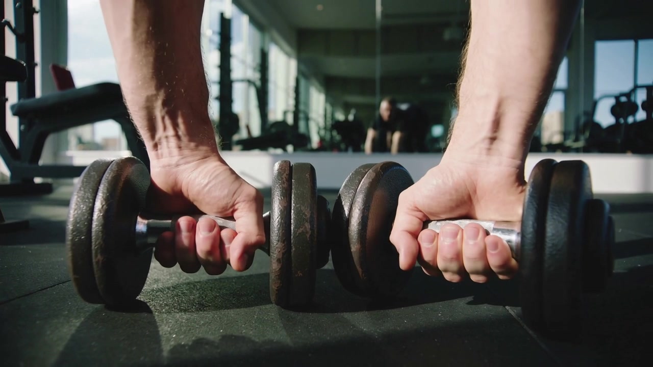 Reaching male lifter wearing short sleeves gripping knurled dumbbells on gym mat, preparing for set