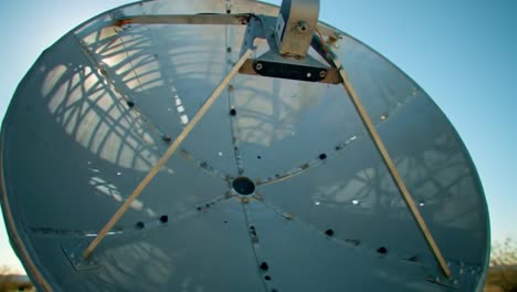 an expansive satellite dish turns to receive signals in a remote desert setting. bright sunlight shines behind the structure, highlighting its intricate design and technology.