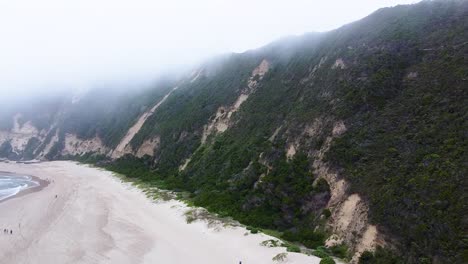 Toma-Aérea-De-Drones-De-La-Playa-De-Sedgefield-Rodeada-De-Dunas-De-Arena-Fértiles-Cubiertas-De-Niebla-De-Nubes-Bajas-Colgantes,-Debajo-De-La-Gente-Que-Pasa-El-Día-Jugando-En-La-Arena,-Cabo-Occidental,-Sudáfrica