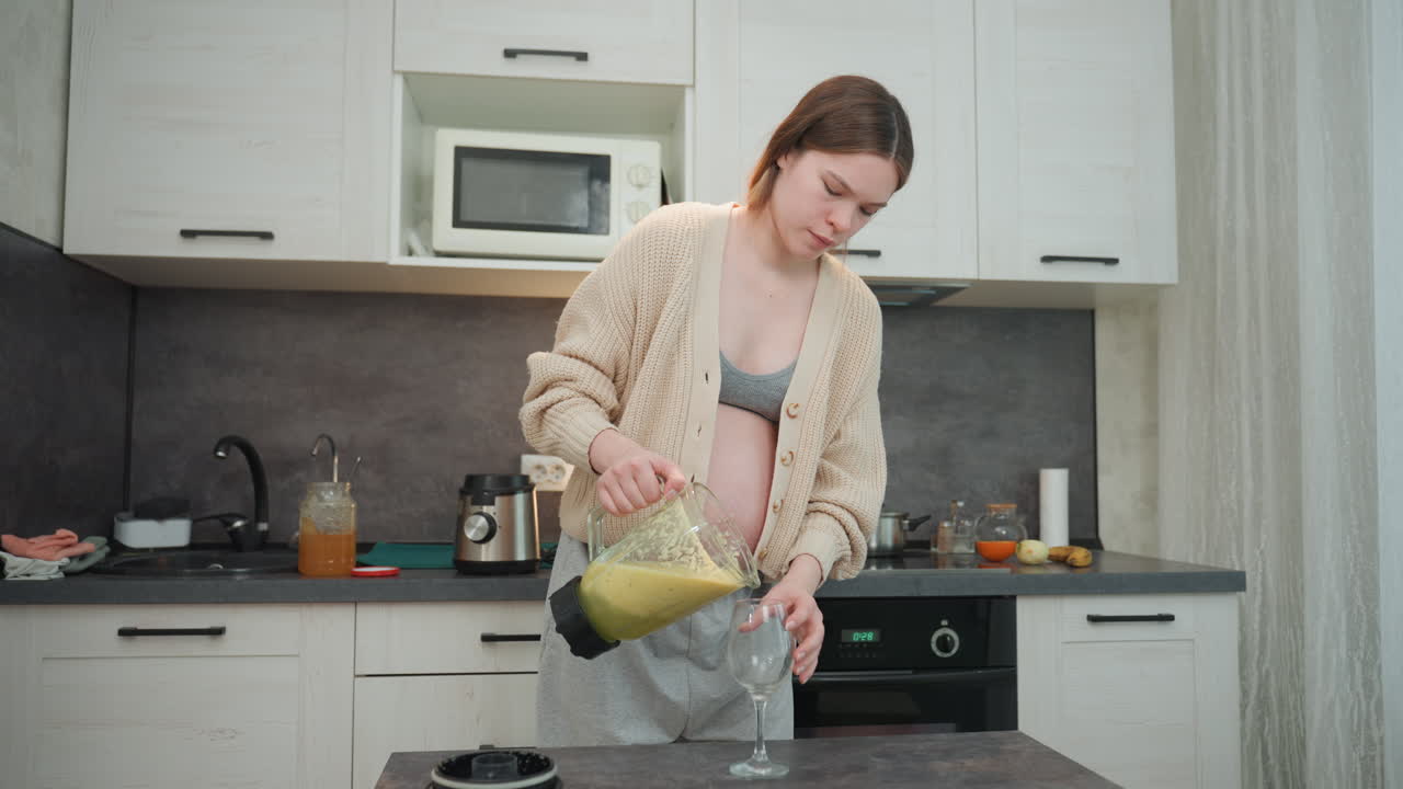 Donna incinta che versa smoothie verde in un bicchiere sul bancone, presa salda della brocca e versamento attento per evitare fuoriuscite, bilancia il bicchiere mentre sorride, rituale di cura di sé e momento nutriente, atmosfera calda e accogliente