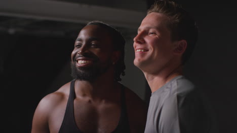 retrato de estudio de amigos masculinos atléticos sonrientes en ropa de fitness entrenando puños golpeando en el gimnasio con iluminación de bajo nivel