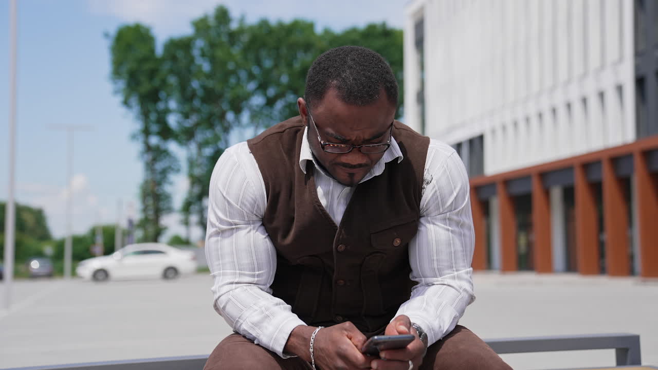 Serious black skinned man looking into phone. Portrait of african american guy with muscular body in suit sitting outdoors and using mobile phone.