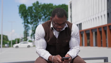 serious black skinned man looking into phone. portrait of african american guy with muscular body in suit sitting outdoors and using mobile phone.