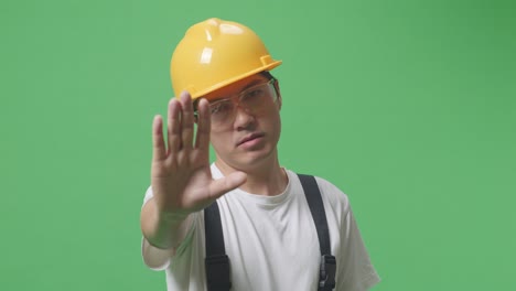 close up de hombre asiático trabajador con gafas de protección y casco de seguridad mirando a la cámara y desaprobando sin señal de mano mientras está de pie en el estudio de fondo de la pantalla verde