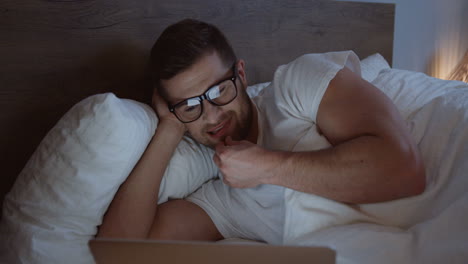 vista de cerca de un hombre sonriente usando una laptop y sonriendo mientras ve algo por la noche en su cama