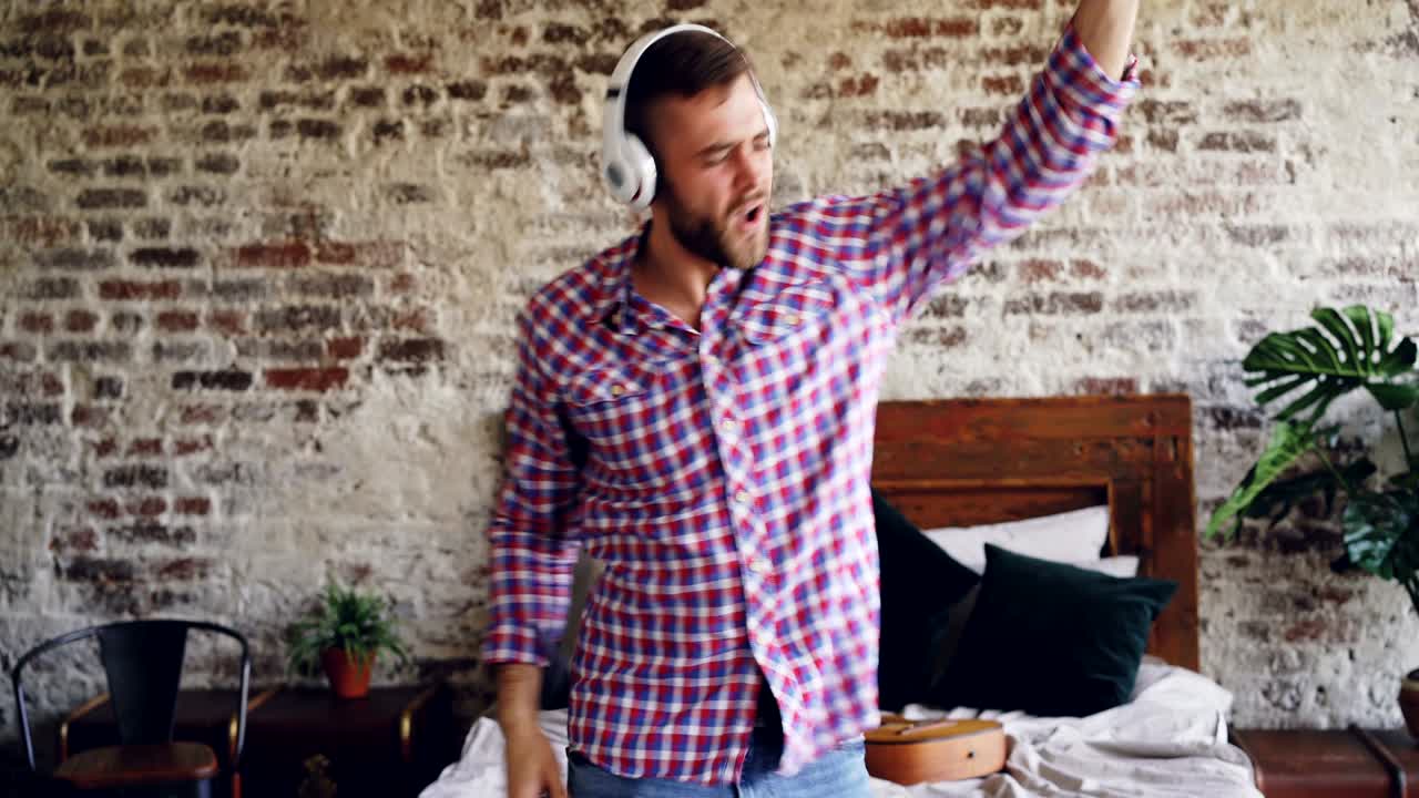 Careless bearded man is dancing and gesturing while listening to music through wireless headphones in bedroom. Excited guy is having fun relaxing in free time.
