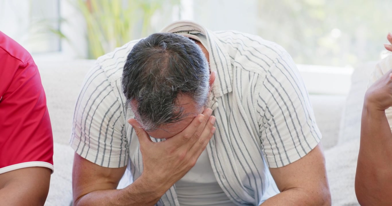 Amici di diverse etnie su un divano chiaro a casa che guardano un uomo sui 40 anni sospirare, coprirsi il viso e condividere i suoi sentimenti.