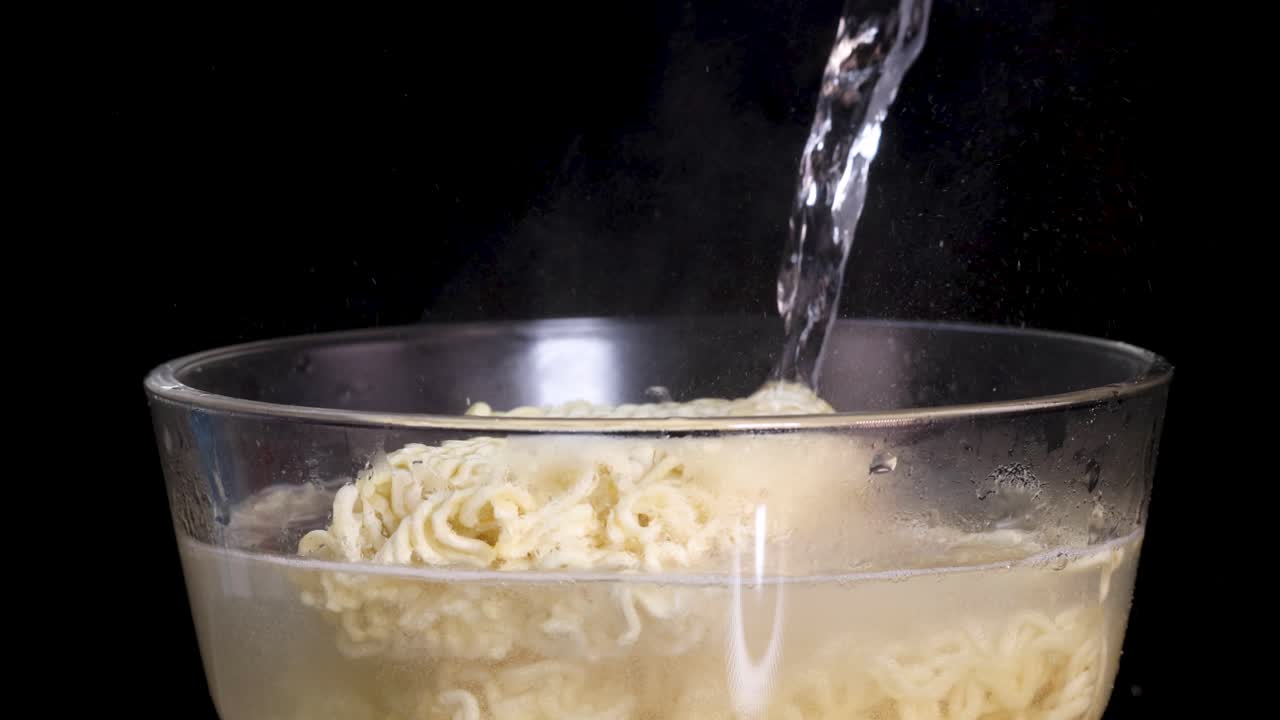 Hot water is poured onto instant noodles in a transparent glass bowl, with steam rising against a dark background. Side view, steady camera, dramatic lighting