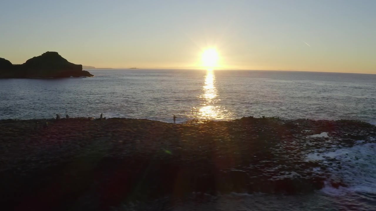 Plano de establecimiento con un paneo aéreo desde la Calzada del Gigante hasta el océano, mostrando un atardecer impresionante con destellos de colores.