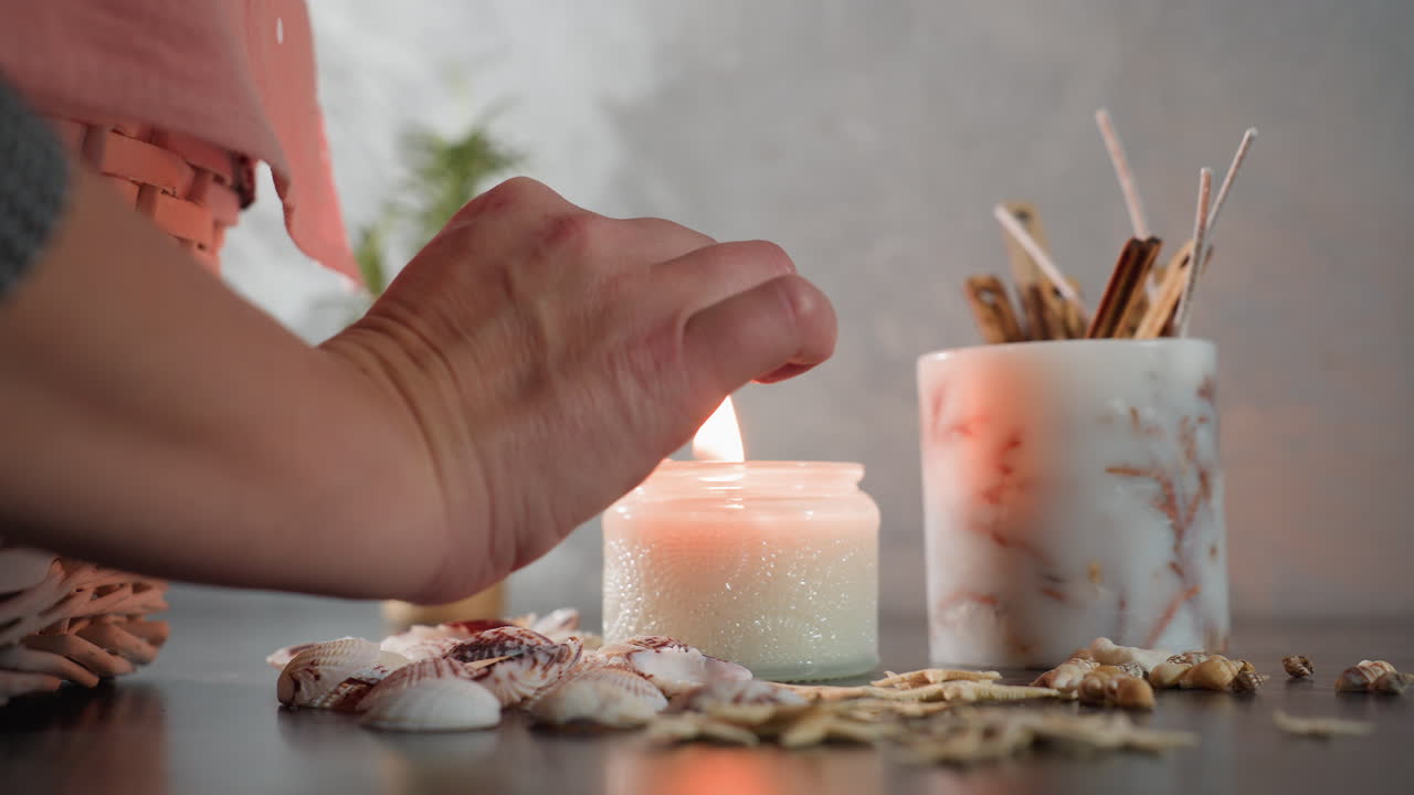 Hand lights candle using matchstick on dark marble surface adorned with decorative seashells, starfish, and snail shells, creating warm cozy ambiance with soft glow