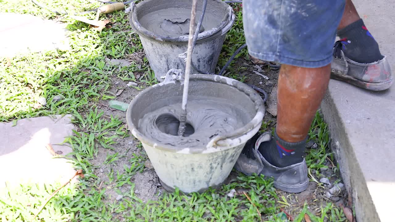 A person mixes cement in a bucket outdoors in Phuket, Thailand, under bright daylight, showcasing manual labor