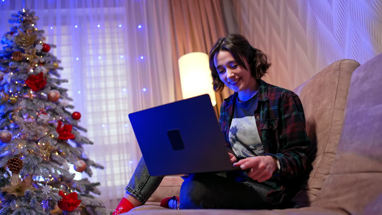 Joyful Caucasian girl looks at the laptop laughing. Low angle view at the lady using a gadget. Christmas season time-spending.