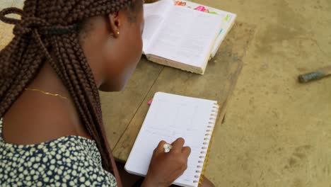 Writing-down-some-notes-while-studying,-a-young-woman-is-educating-herself-in-her-own-home-in-a-village-in-Kumasi,-Ghana-in-Africa