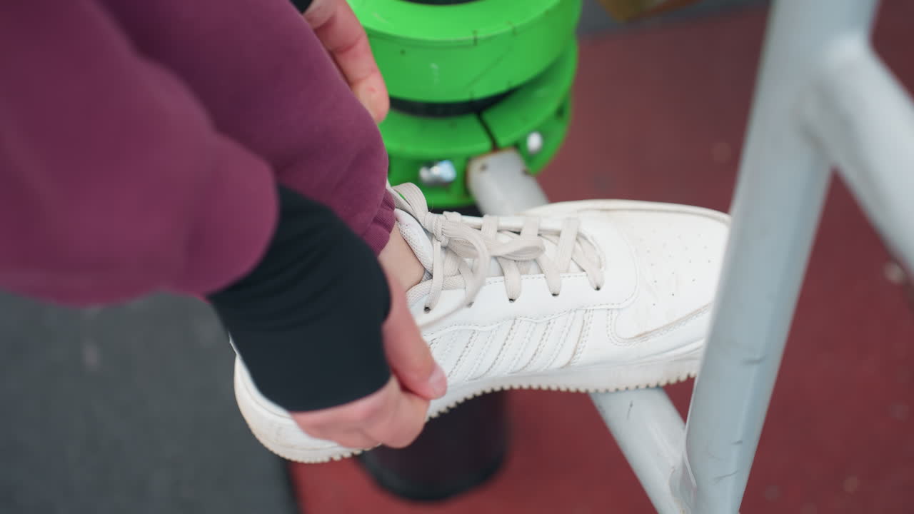 High angle view of female coach tying canvas sneaker lace on iron exercise bar in urban outdoor fitness park before strength training session near modern apartment building under open sky