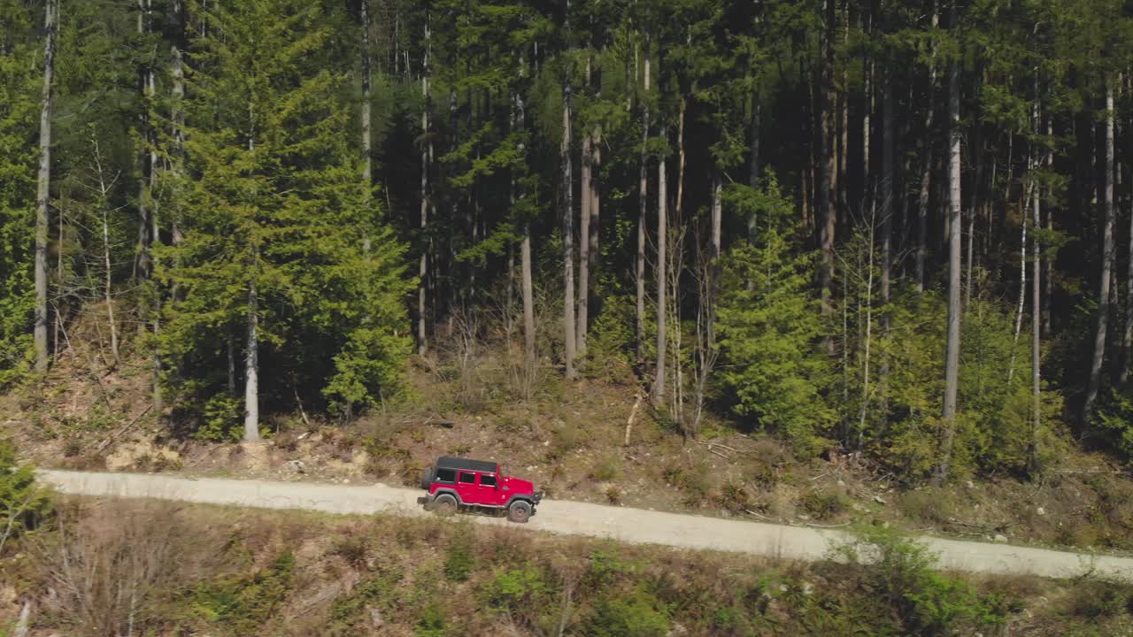 SUV Jeep rosso fuoristrada che guida su una strada sterrata forestale, avvicinandosi e superando una fitta foresta verde sullo sfondo. Vista aerea laterale che segue il veicolo mentre si sposta a destra e accelera.