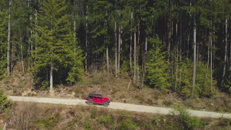 red jeep suv off road vehicle driving on dirt logging road approaching and passing dense green forest view in background aerial beside trucking tracking as truck moves to right accelerating passed