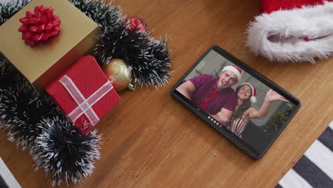 pareja caucásica sonriente con sombreros de santa en una videollamada de navidad en una tableta