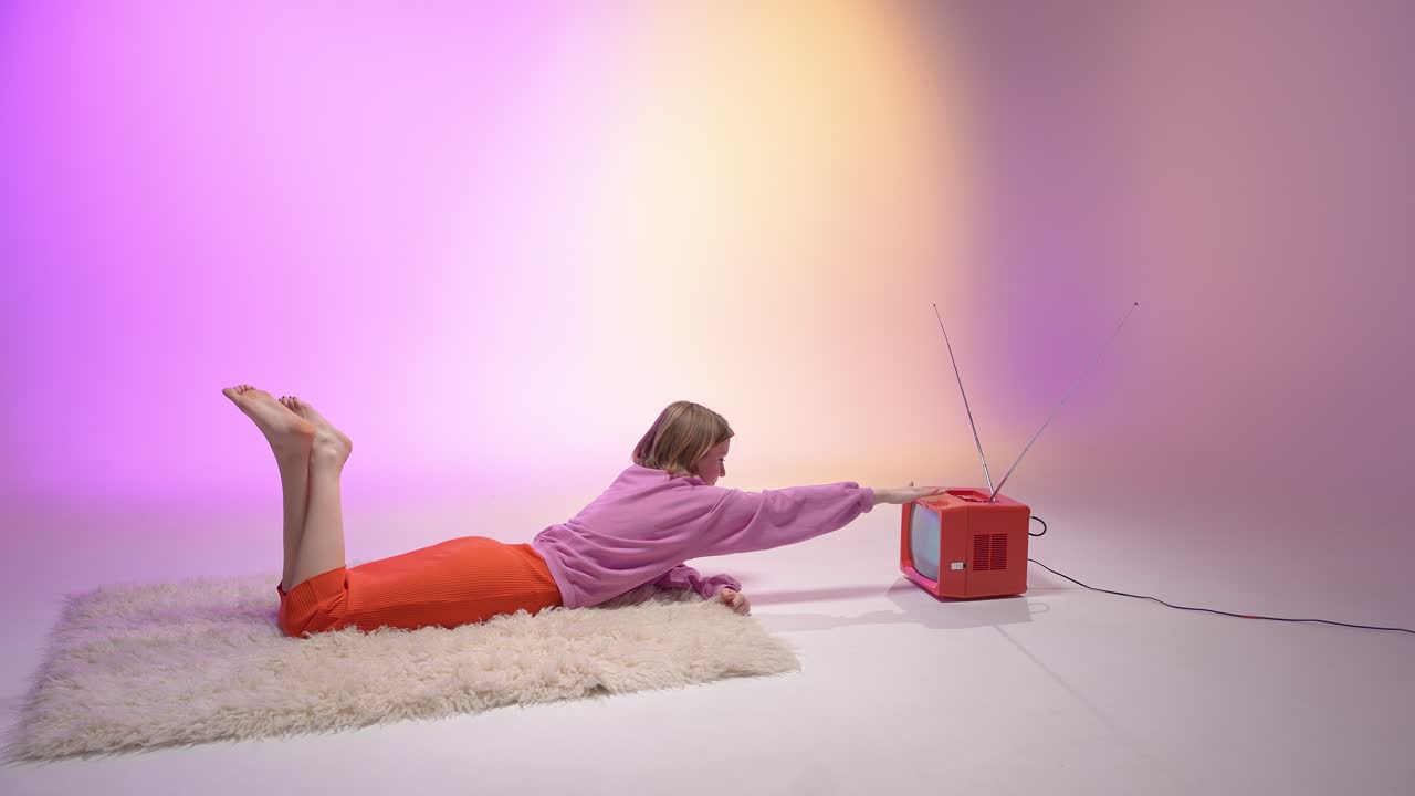 A young woman lies on a fluffy rug and reaches toward a vintage red television. The background glows with pink and yellow hues, blending nostalgic aesthetics with a cozy, retro-inspired vibe