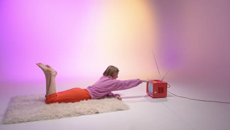 a young woman lies on a fluffy rug and reaches toward a vintage red television. the background glows with pink and yellow hues, blending nostalgic aesthetics with a cozy, retro-inspired vibe